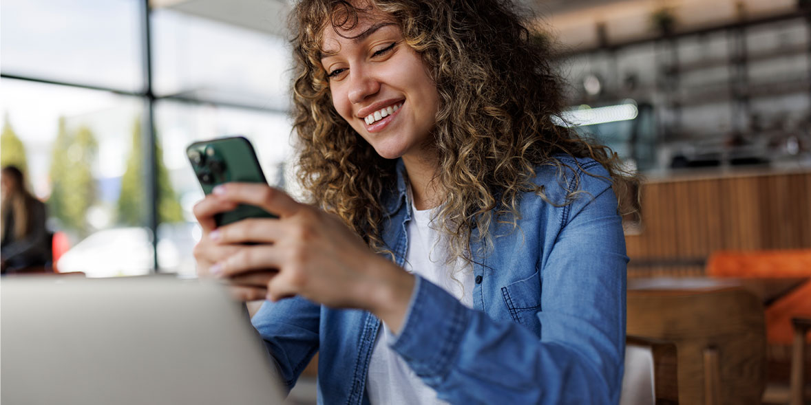 Smiling young woman with curly hair sits at a cafe, holding a smartphone while looking at her laptop.