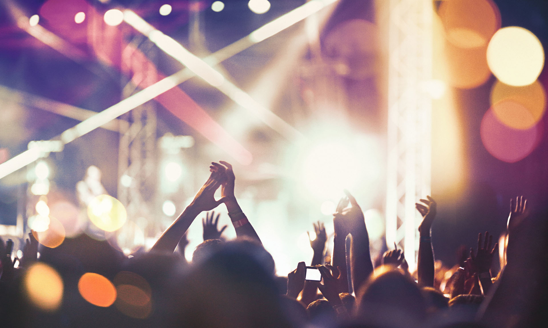 Crowd with raised hands at a concert, vibrant lights in the background creating a festive atmosphere.